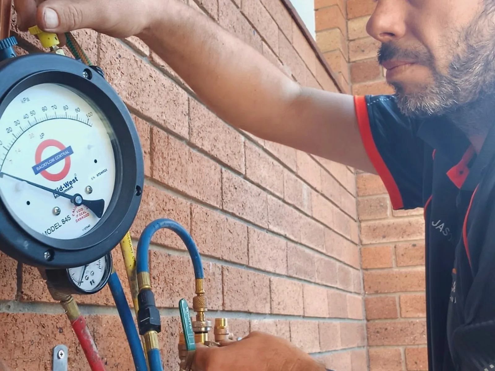 Jason Breingan of Flowtek Services testing a backflow prevention device on a brick wall, a differential pressure gauge and red, blue and yellow test hoses connected to the device