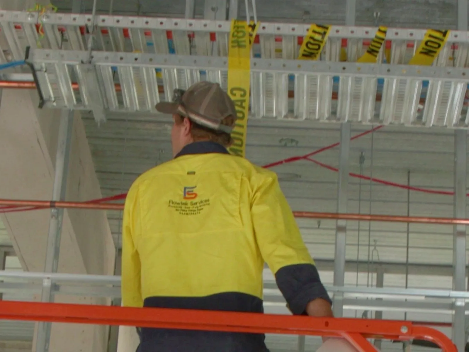 A Flowtek Services plumber in a yellow hi-vis shirt working from an elevated work platform on services pipework in a high commercial ceiling
