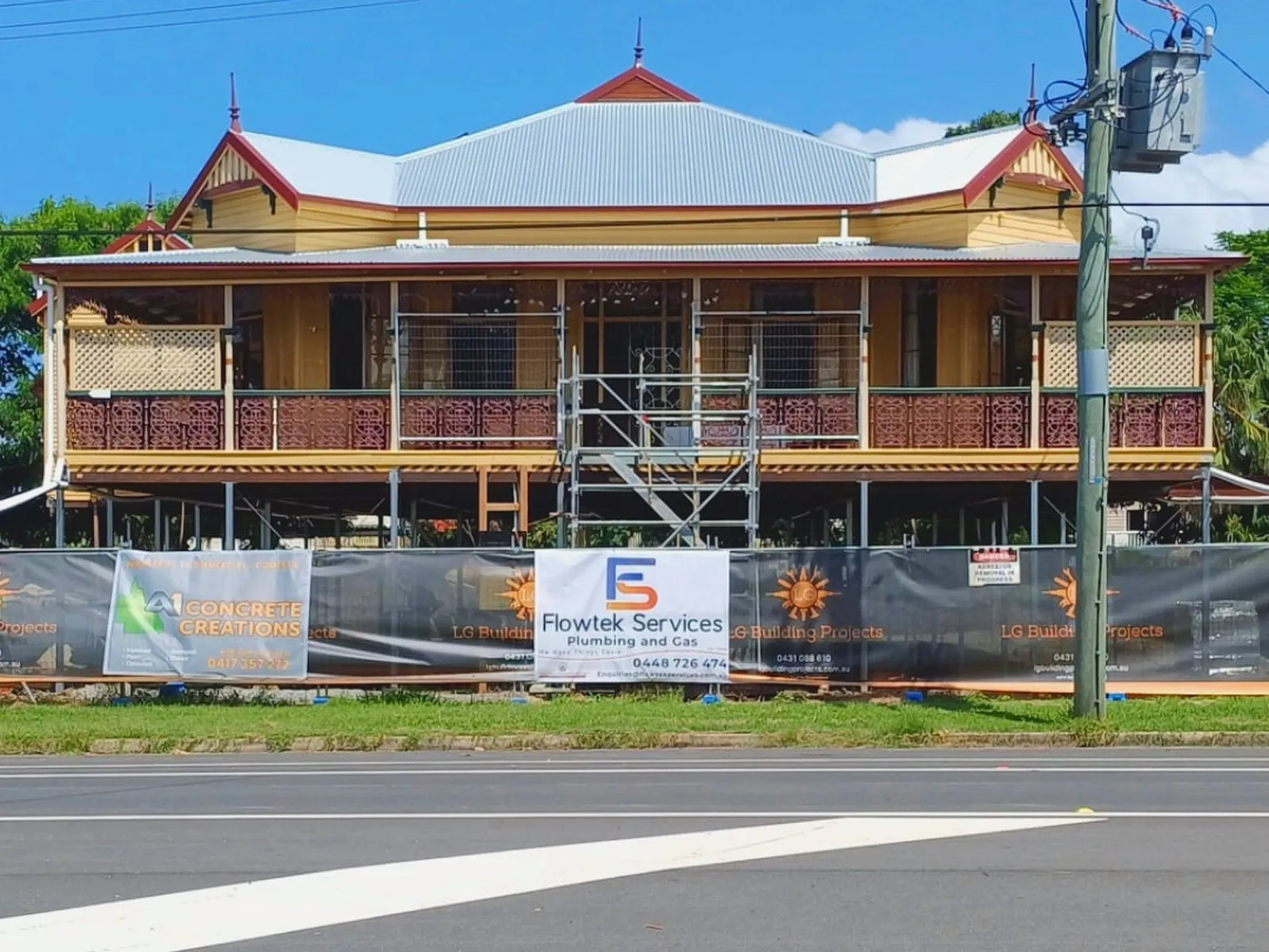 A large two-storey heritage Queenslander under restoration behind site fencing, with a Flowtek Services Plumbing and Gas banner on the hoarding