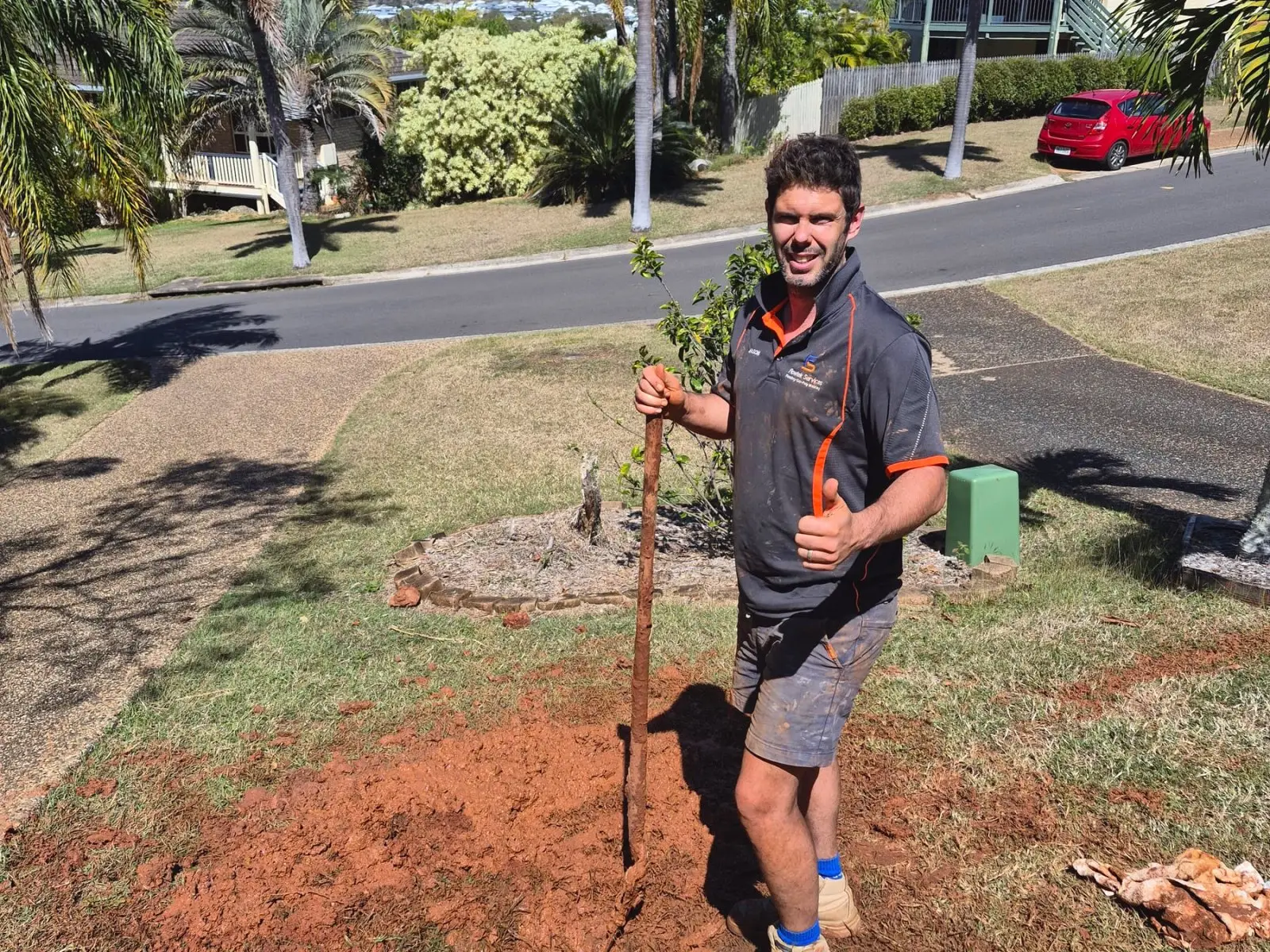 Jason Breingan of Flowtek Services laughing, leaning on a shovel beside an open trench on a suburban verge with palms and a Queenslander house behind him