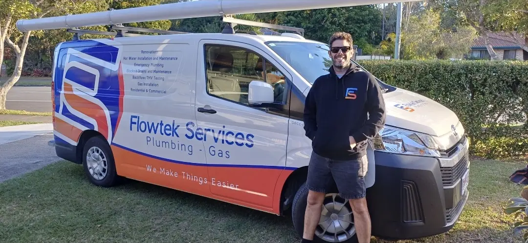 Jason Breingan of Flowtek Services standing with his arms folded in front of the white Flowtek work ute in Yeppoon, wearing the charcoal polo with the orange collar, ladders on the rack behind him