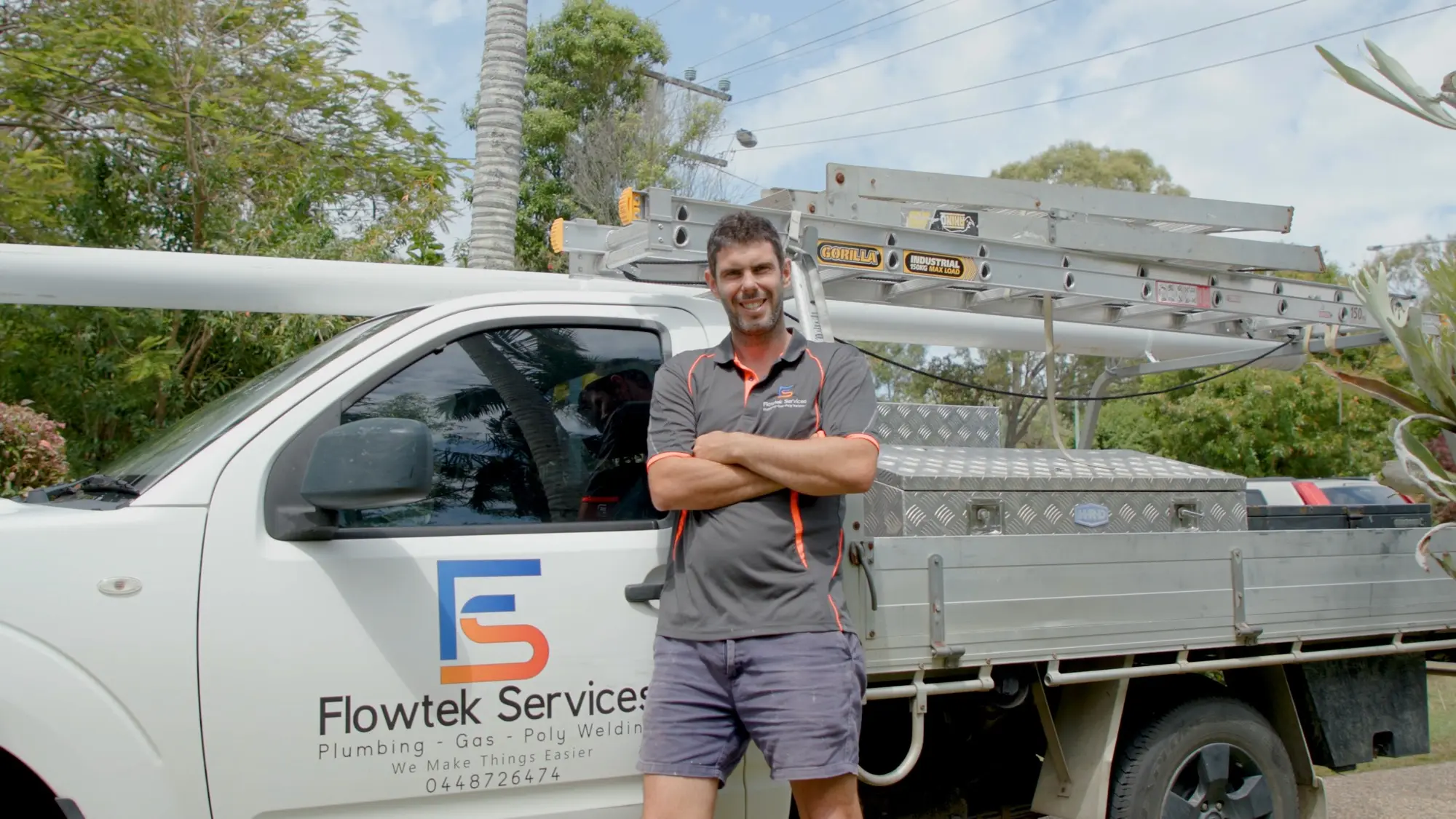 Jason Breingan of Flowtek Services standing with his arms folded in front of the white Flowtek work ute in Yeppoon, wearing the charcoal polo with the orange collar, ladders on the rack behind him