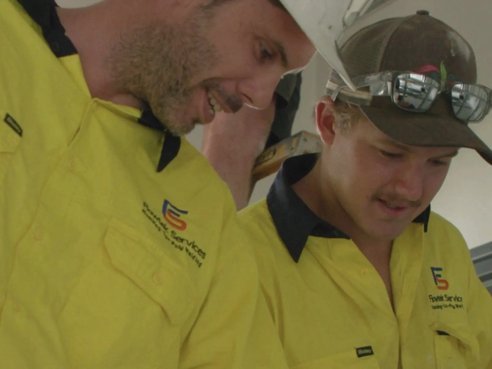 Two Flowtek Services plumbers in yellow hi-vis shirts on a commercial site, heads together over a detail, working it out between them