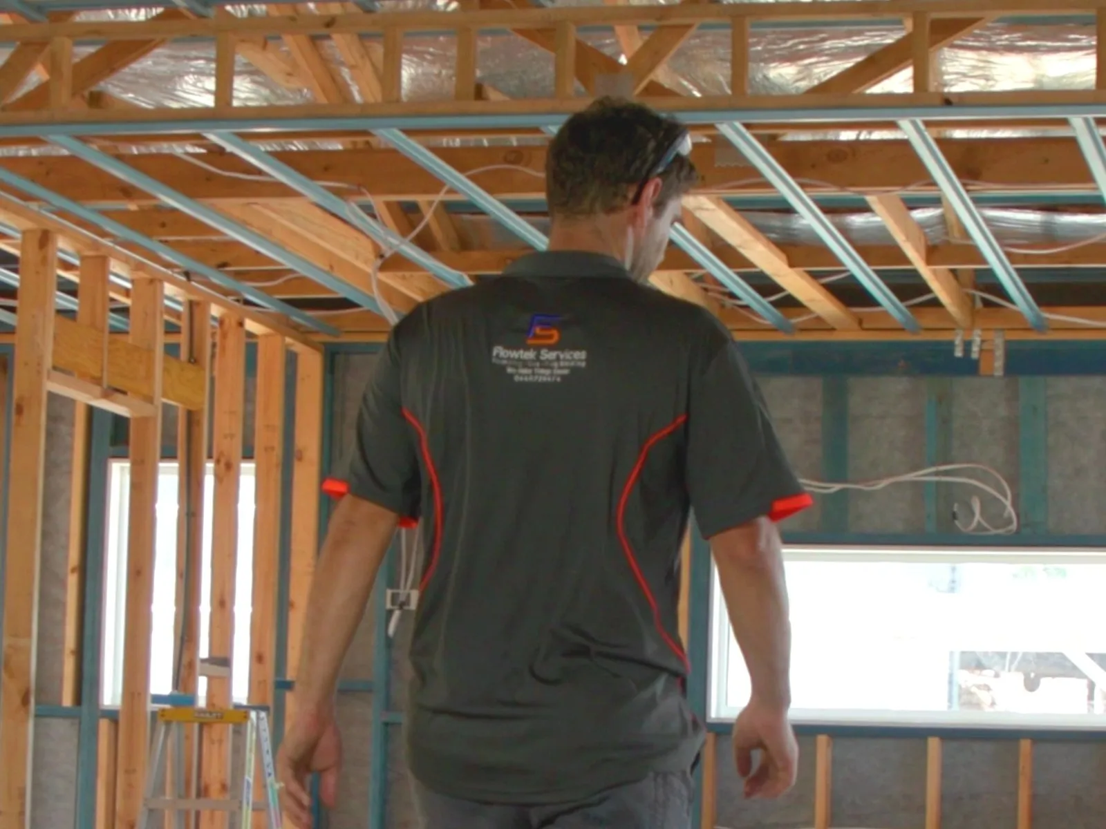 A Flowtek Services plumber walking through a house under construction, exposed pine timber frame and roof trusses, teal steel window frames and a raw concrete floor