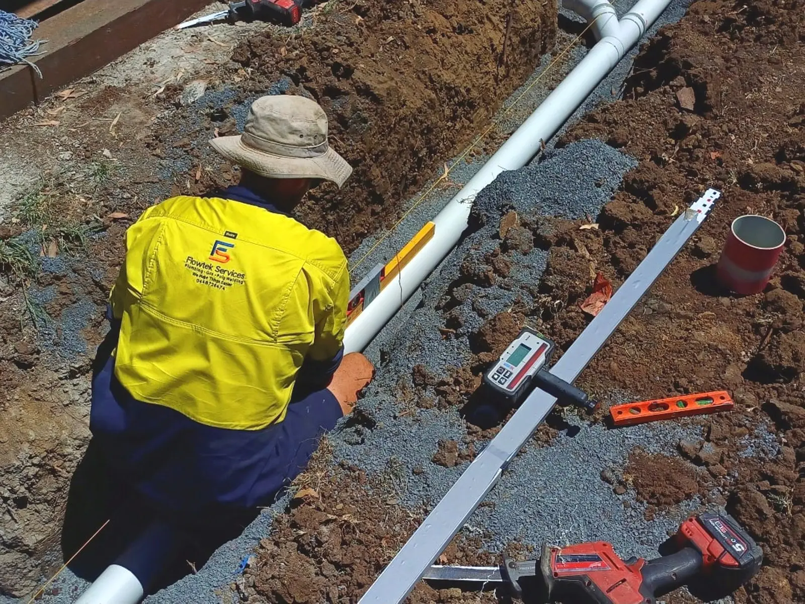 A Flowtek Services plumber in a hi-vis Flowtek shirt laying PVC drainage pipe into a gravel-bedded trench, with a laser level, spirit level and reciprocating saw beside him