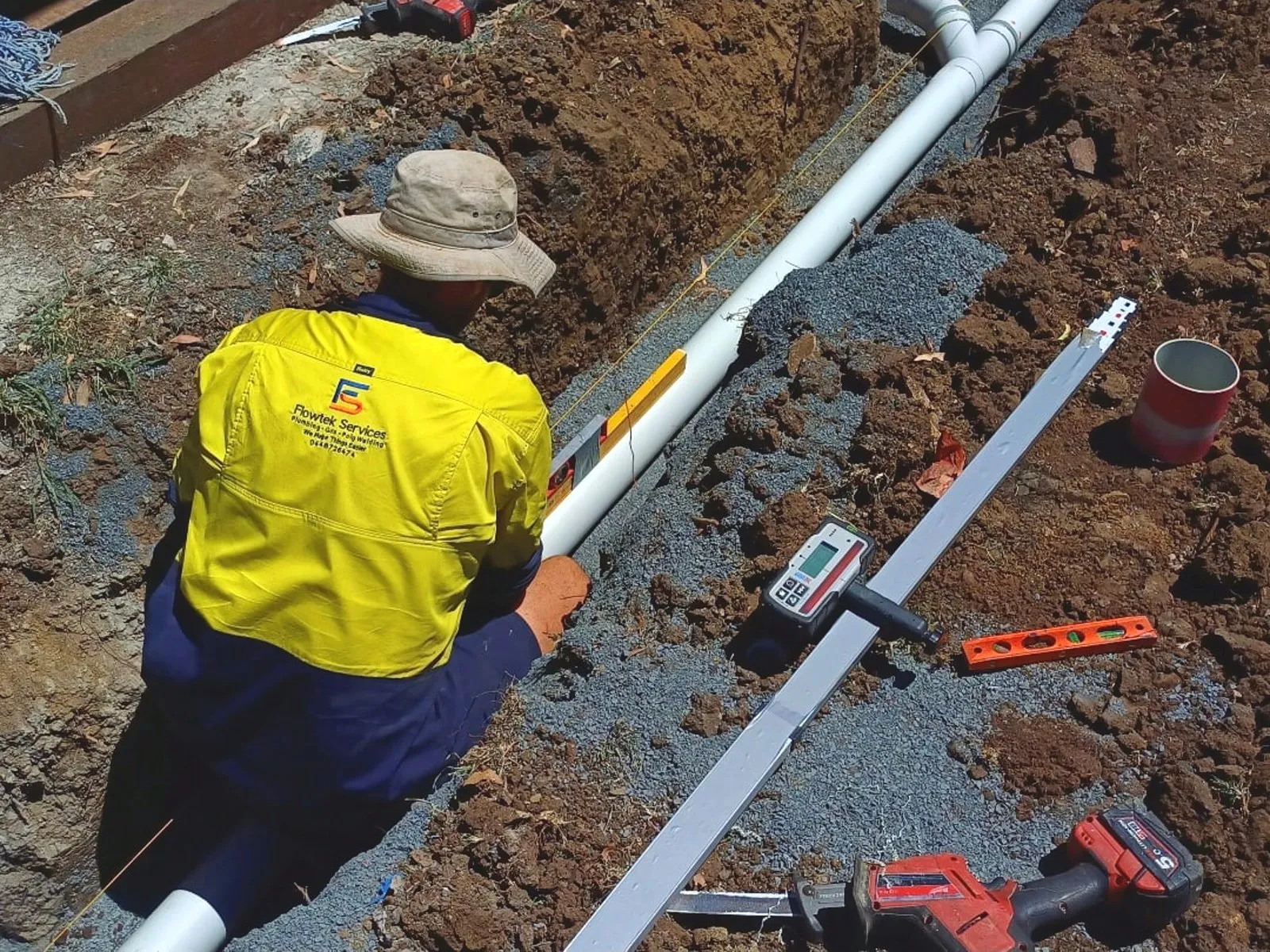 A Flowtek Services plumber in a hi-vis Flowtek shirt laying PVC drainage pipe into a gravel-bedded trench, with a laser level, spirit level and reciprocating saw beside him