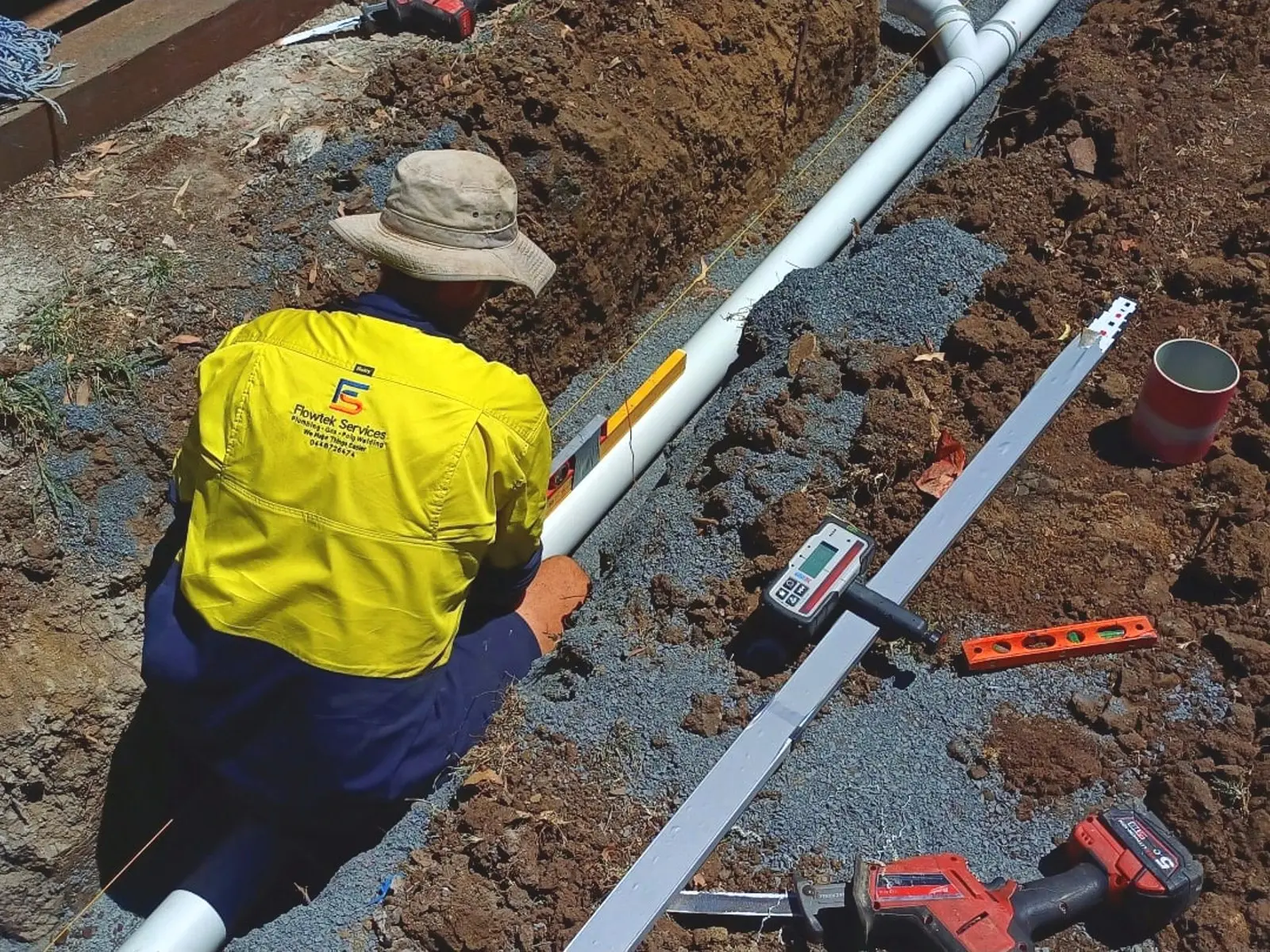 A Flowtek Services plumber in a hi-vis Flowtek shirt laying PVC drainage pipe into a gravel-bedded trench, with a laser level, spirit level and reciprocating saw beside him