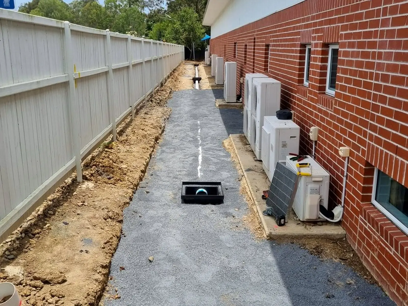 A gravel drainage path running beside a red brick building at Taranganba, with a black drainage pit set into the gravel and an open trench cut along the fence line