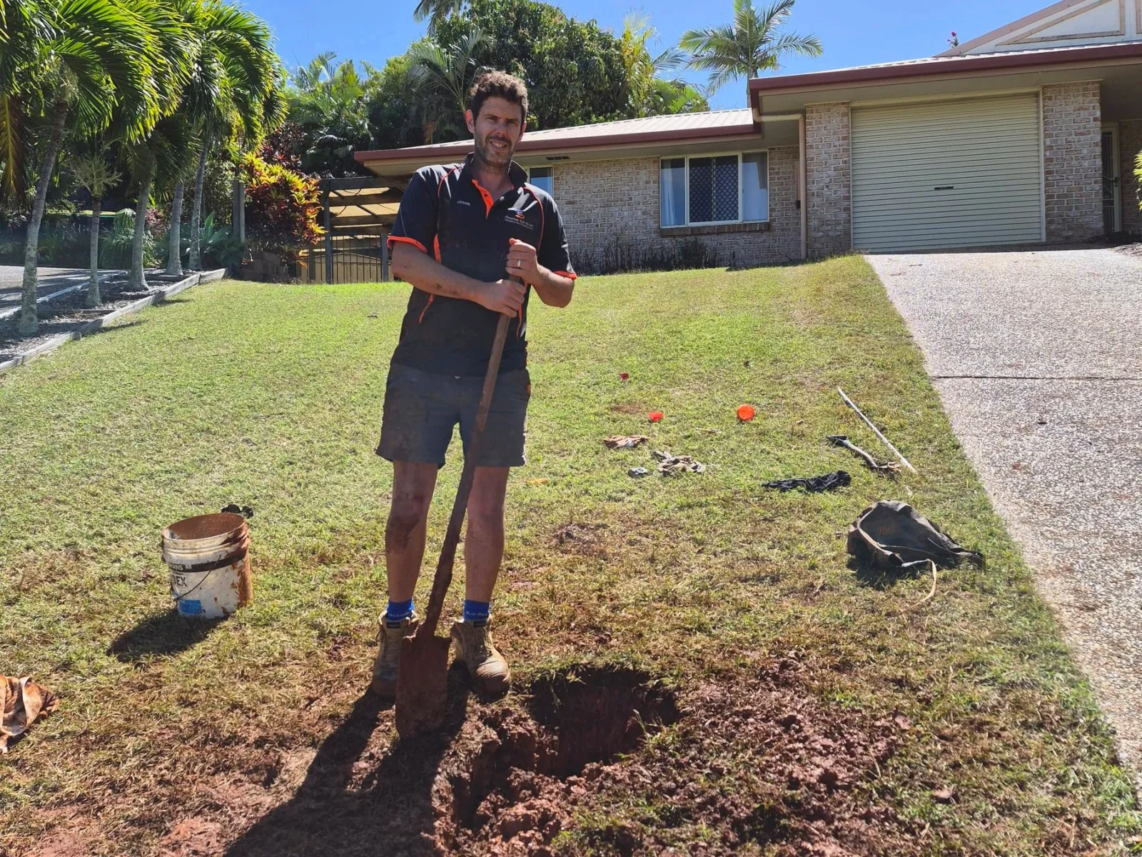 Jason Breingan of Flowtek Services standing in a suburban front yard beside a freshly dug trench with a shovel, in hard Central Queensland sun